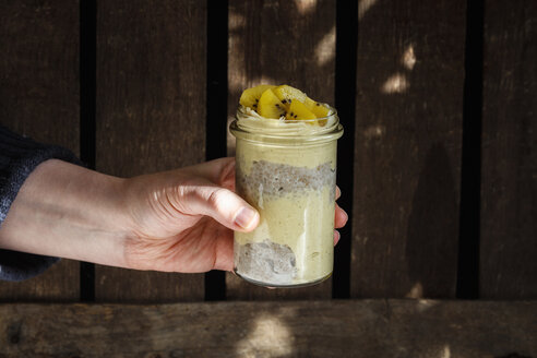 Männerhand hält Glas Chia-Pudding mit Mango-Smoothie garniert mit Kiwi - EVGF03356