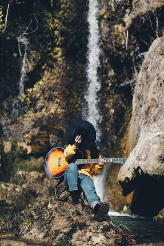 Spanien, Lleida, junge Frau mit Gitarre sitzt auf einem Felsen vor einem Wasserfall und bedeckt ihr Gesicht mit einem Hut, lizenzfreies Stockfoto