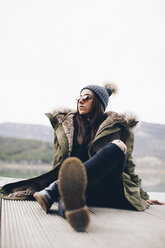 Portrait of young woman sitting on jetty wearing sunglasses and bobble hat - OCAF00197