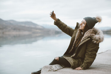 Lächelnde junge Frau sitzt auf dem Steg und macht ein Selfie mit ihrem Smartphone - OCAF00201