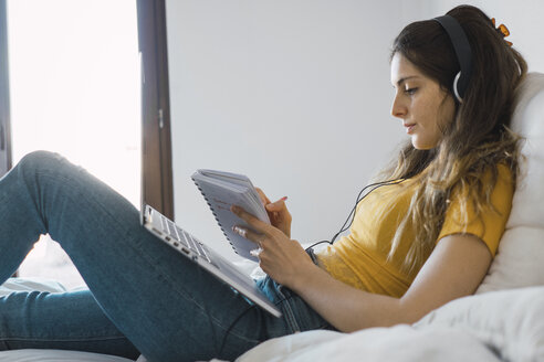 Young woman with laptop and headphones lying on bed writing down something - KKAF00971