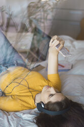 Junge Frau mit Kopfhörern liegt auf dem Bett und macht ein Selfie mit ihrem Smartphone - KKAF00975