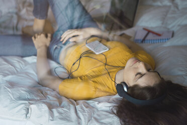 Young woman lying on bed listening music with headphones and smartphone - KKAF00976