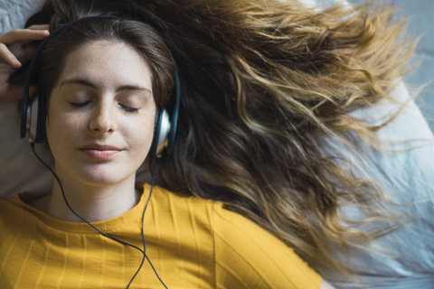 Porträt einer lächelnden jungen Frau, die auf dem Bett liegt und mit Kopfhörern Musik hört, lizenzfreies Stockfoto