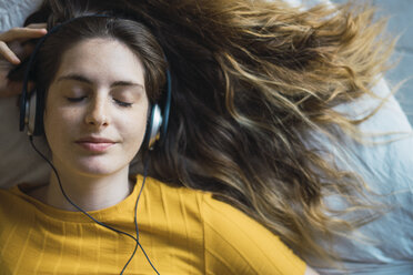 Portrait of smiling young woman lying on bed listening music with headphones - KKAF00978