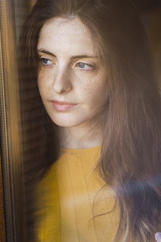 Porträt einer nachdenklichen jungen Frau mit langen braunen Haaren, die aus dem Fenster schaut, lizenzfreies Stockfoto