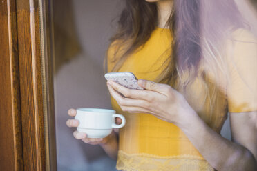 Young woman with coffee cup and cell phone standing behind windowpane, close-up - KKAF00982