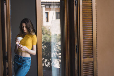 Junge Frau mit Kaffeetasse, die sich gegen ein Türgehäuse lehnt und auf ihr Handy schaut - KKAF00983