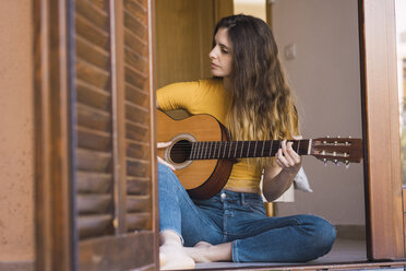 Young woman sitting on the floor at home playing guitar - KKAF00984