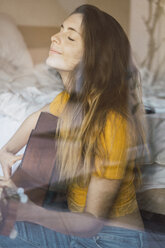 Portrait of content young woman sitting behind windowpane playing guitar - KKAF00985