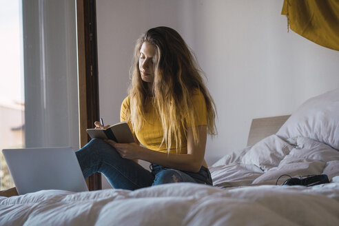 Junge Frau mit Laptop sitzt auf dem Bett und schreibt etwas auf - KKAF00987