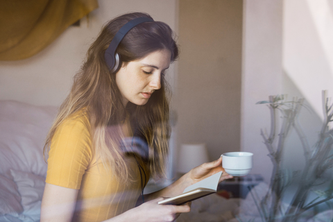 Junge Frau mit Kaffeetasse, Kopfhörern und Notizbuch hinter einer Fensterscheibe, lizenzfreies Stockfoto