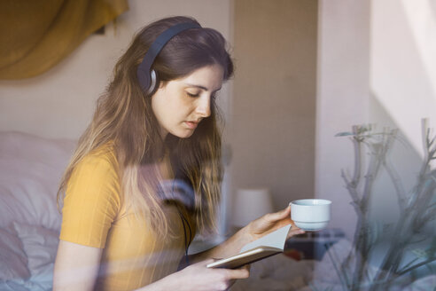 Junge Frau mit Kaffeetasse, Kopfhörern und Notizbuch hinter einer Fensterscheibe - KKAF00988