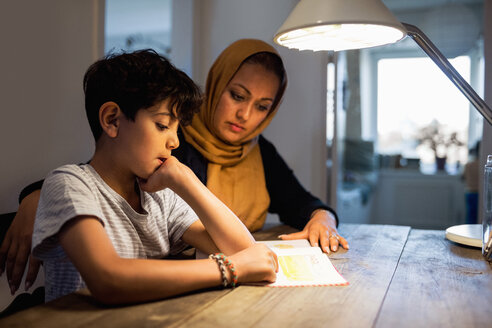 Mother and son reading book under illuminated desk lamp at home - MASF02981