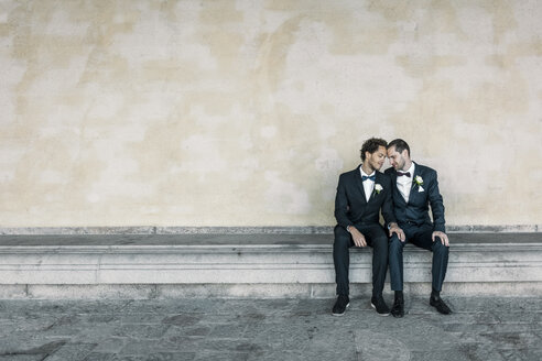 Newlywed gay couple sitting on bench against wall - MASF02991