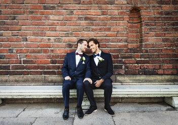 Neuvermähltes homosexuelles Paar sitzt auf einer Bank vor einer Backsteinmauer - MASF02992