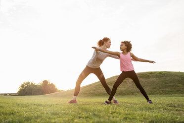 Mother assisting daughter practicing exercise on grass at park against sky - MASF02998