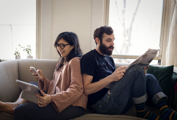 Frau benutzt Mobiltelefon, während ihr Mann auf dem Sofa Zeitung liest - CAVF36352