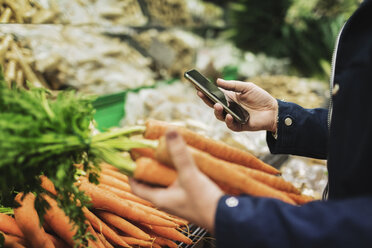 Midsection of woman using phone while buying carrots in supermarket - MASF03006