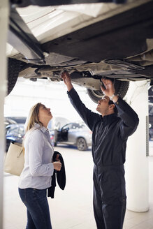 Mechanic with female customer standing under car at repair shop - MASF03014