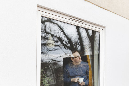 Mann hält Kaffeetasse und schaut durch ein Fenster, während er in einem Haus steht - MASF03016