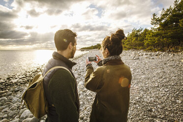 Mann sieht Frau an, die mit ihrem Handy am Strand das Meer gegen den Himmel fotografiert - MASF03033