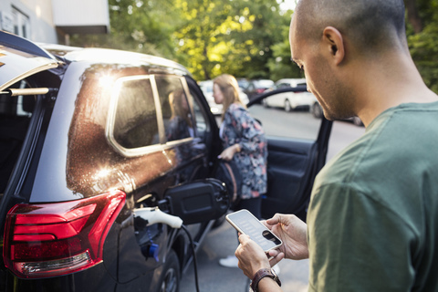 Mann benutzt Smartphone mit Frau, die im Hintergrund am Auto steht, lizenzfreies Stockfoto