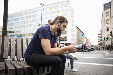 Mann hört Musik über sein Smartphone, während er neben einer Frau auf einer Holzbank in der Stadt sitzt - MASF03042