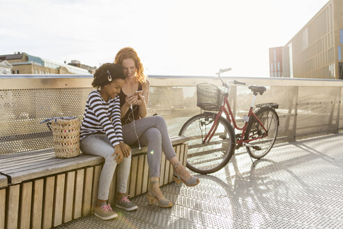 Happy girl sitting with mother using mobile phone while sitting on bench against sky - MASF03067