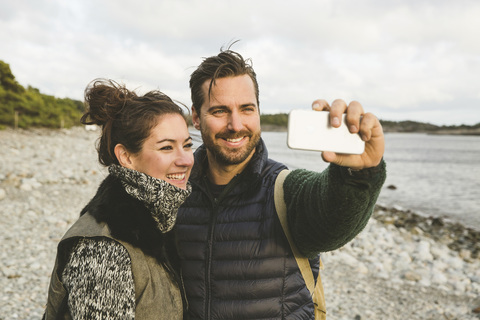 Glückliches Paar nimmt Selfie am Strand gegen den Himmel bei Sonnenuntergang, lizenzfreies Stockfoto