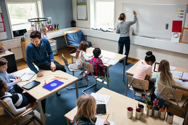 Blick von oben auf eine Lehrerin, die an die Tafel schreibt, während ein männlicher Pädagoge mit den Schülern am Schreibtisch im Klassenzimmer sitzt - MASF03097