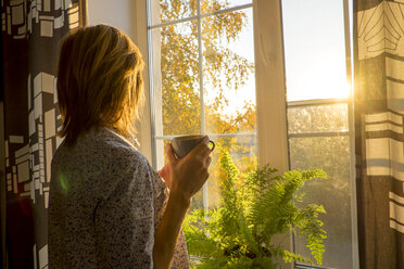 Seitenansicht einer Frau, die eine Kaffeetasse hält, während sie zu Hause am Fenster steht - CAVF36475
