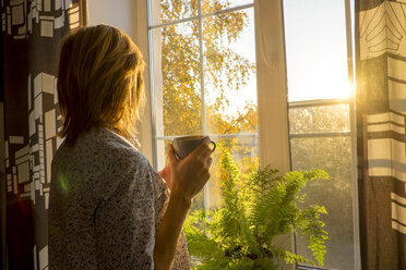 Side view of woman holding coffee cup while standing by window at home - CAVF36475