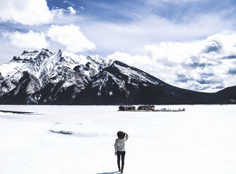 Rear view of woman looking at view while standing by frozen Lake Minnewanka against cloudy sky - CAVF36512