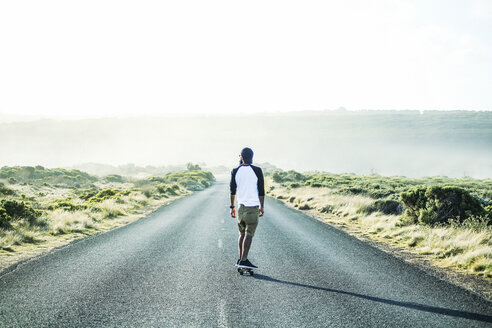 Rear view of man skateboarding on country road against clear sky - CAVF36520