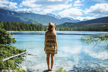 Rückansicht einer Frau mit Hut, die die Aussicht betrachtet, während sie auf einem Felsen am See im Jasper National Park steht - CAVF36542