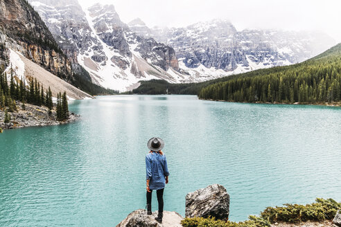 Rückansicht einer Frau mit Hut, die auf einem Felsen am Moraine Lake im Banff National Park steht und die Aussicht betrachtet - CAVF36547