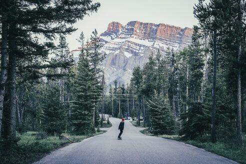 Frau fährt Skateboard auf einer Landstraße zwischen Bäumen und Bergen im Banff National Park - CAVF36561