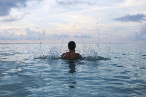 Rückansicht eines Mannes ohne Hemd, der im Meer vor einem bewölkten Himmel Wasser spritzt - CAVF36566
