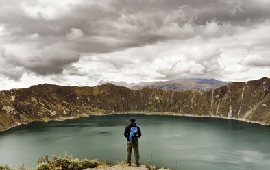 Rear view of man standing on cliff against Quilotoa and cloudy sky - CAVF36599