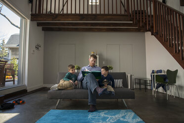 Vater zeigt seinen Söhnen ein Buch, während er zu Hause auf dem Sofa sitzt - CAVF36622