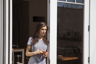 Vorderansicht einer Frau, die eine Kaffeetasse hält und am Fenster steht - CAVF36626