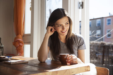 Happy woman holding coffee cup and sitting by table at home - CAVF36629