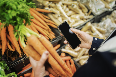 Ausgeschnittenes Bild einer Frau, die beim Kauf von Karotten im Supermarkt telefoniert, lizenzfreies Stockfoto