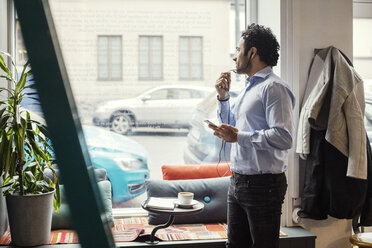 Kreativer Mann benutzt Kopfhörer, während er im Büro am Fenster steht - MASF03118