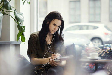 Kreative Geschäftsfrau mit Kopfhörern beim Schreiben in einem Spiralnotizbuch im Büro - MASF03119