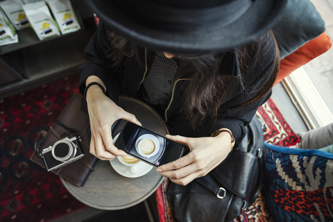 Direkt über der Aufnahme einer jungen Frau, die mit ihrem Smartphone im Kreativbüro Kaffee fotografiert, lizenzfreies Stockfoto