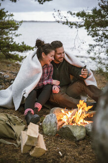 In eine Decke eingewickeltes Paar, das beim Sitzen an einer Feuerstelle auf einem Campingplatz ein Mobiltelefon benutzt - MASF03159