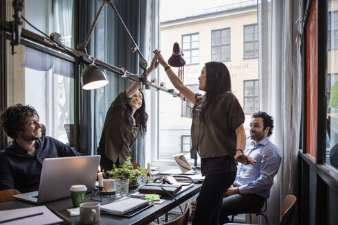 Female colleagues giving high-five while standing at table in meeting - MASF03167