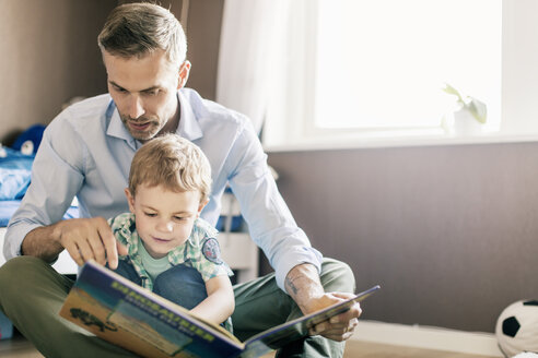 Vater zeigt auf seinen Sohn und liest ihm ein Buch vor, während er zu Hause auf dem Boden sitzt - MASF03174